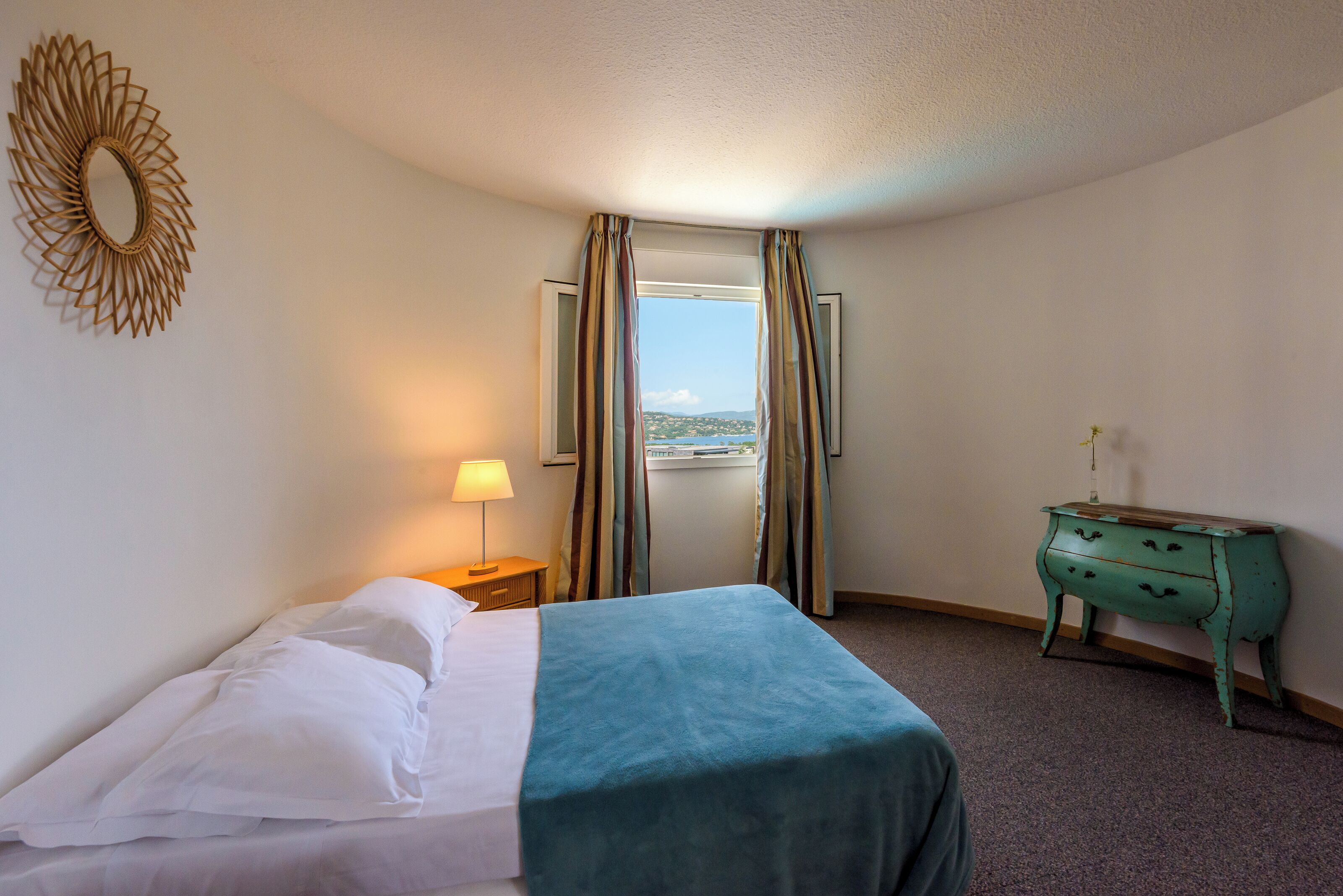 Ein Hotelzimmer mit Bett, Lampe, Kommode und Meerblick durch Vorhänge.