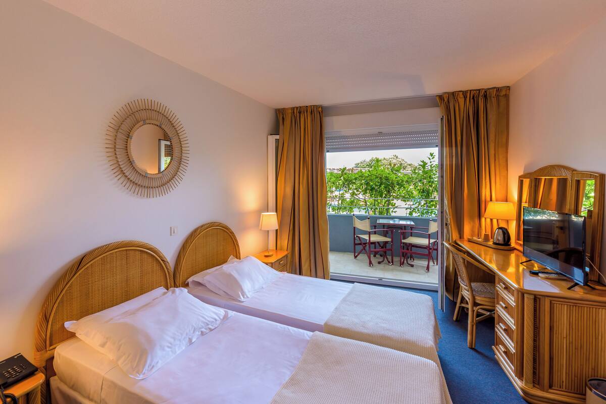 Habitación de hotel con dos camas, televisión, escritorio y balcón con mobiliario exterior.