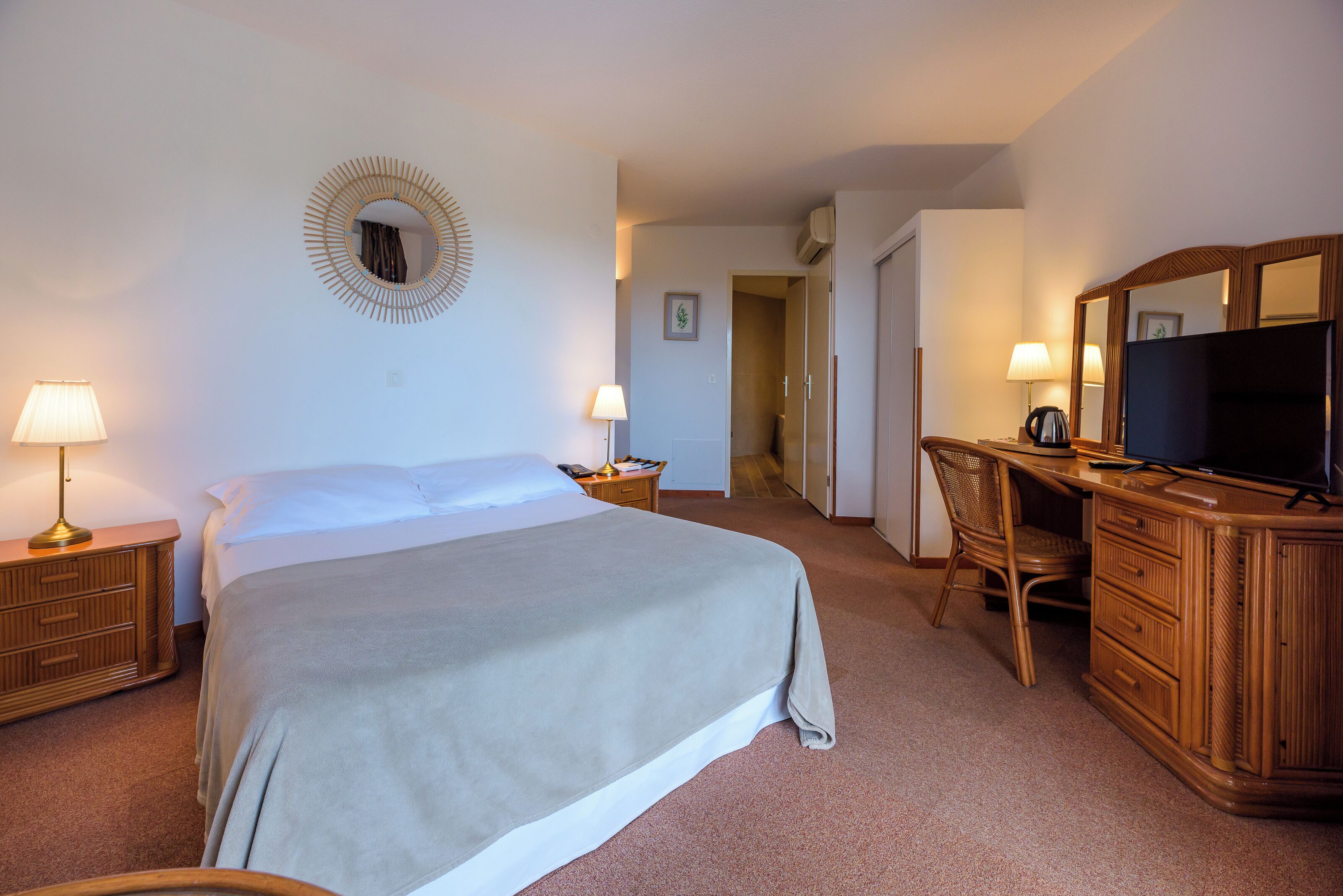 Quarto de hotel com cama, luminárias de cabeceira, televisão, escrivaninha e espelho.