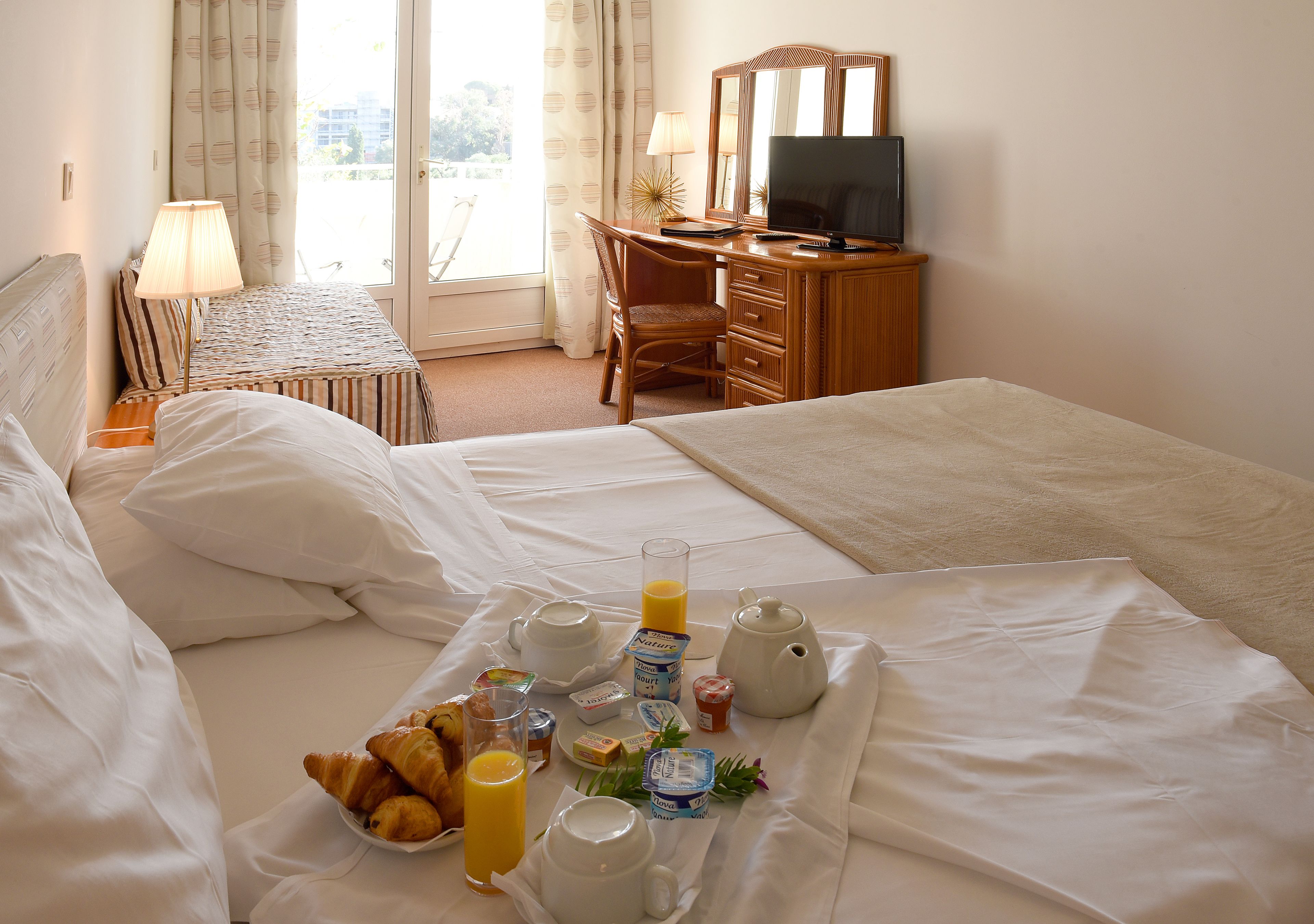 A hotel room with two beds, a desk with a TV, and a breakfast tray with croissants, juice, and coffee.