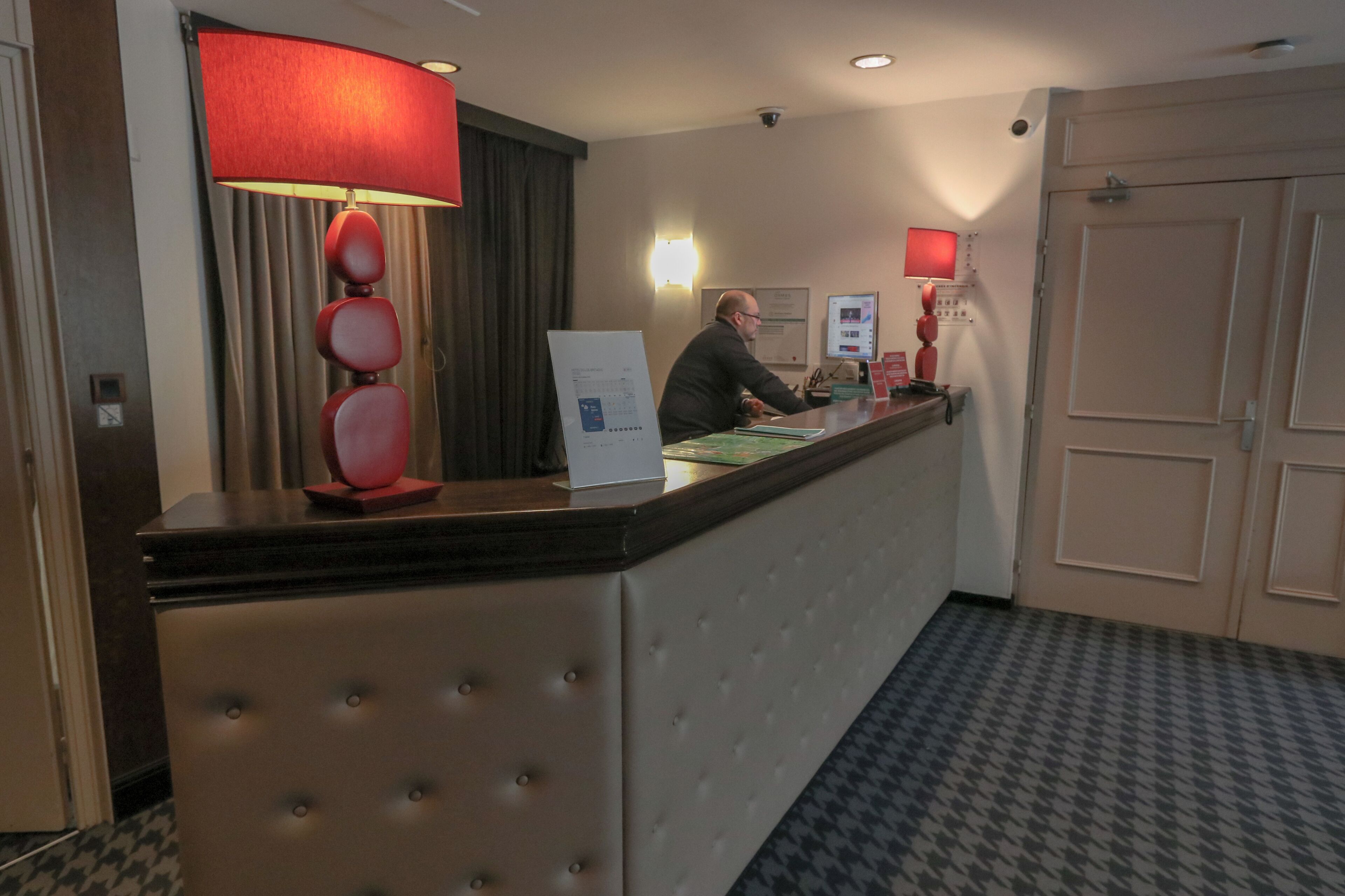 A hotel reception area with a reception desk, a red lamp, a bulletin board, and a wall-mounted security camera.