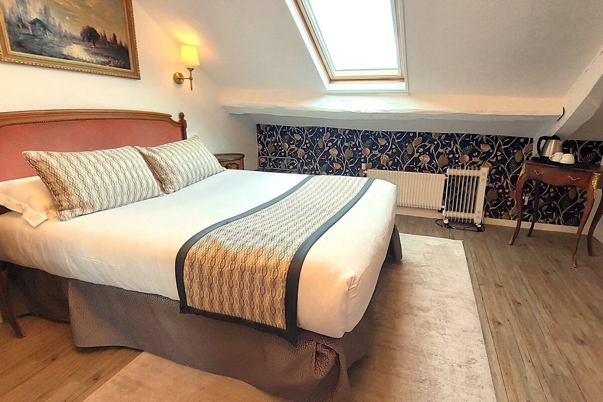 A bedroom with a bed, a radiator, a nightstand, a table, and a painting on the wall.