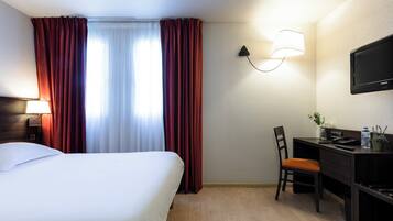 A hotel room with a bed, a desk, a chair, a lamp, a TV, and a window with curtains.