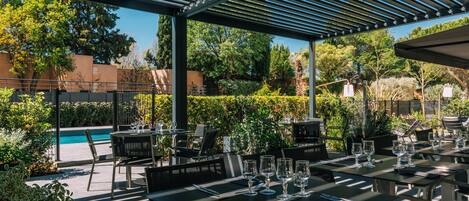 An outdoor dining area with tables and chairs, a pergola, and a pool in the background.