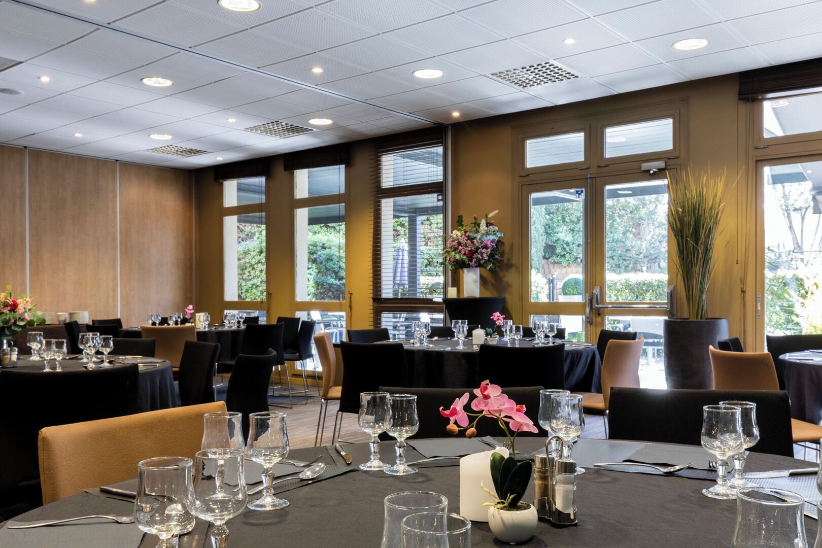 A dining area with tables set for service, large windows, and indoor plants.