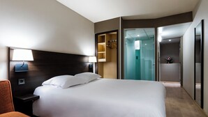 A hotel room with a bed, a nightstand, a lamp, a chair, a wardrobe, and a bathroom with a glass door.