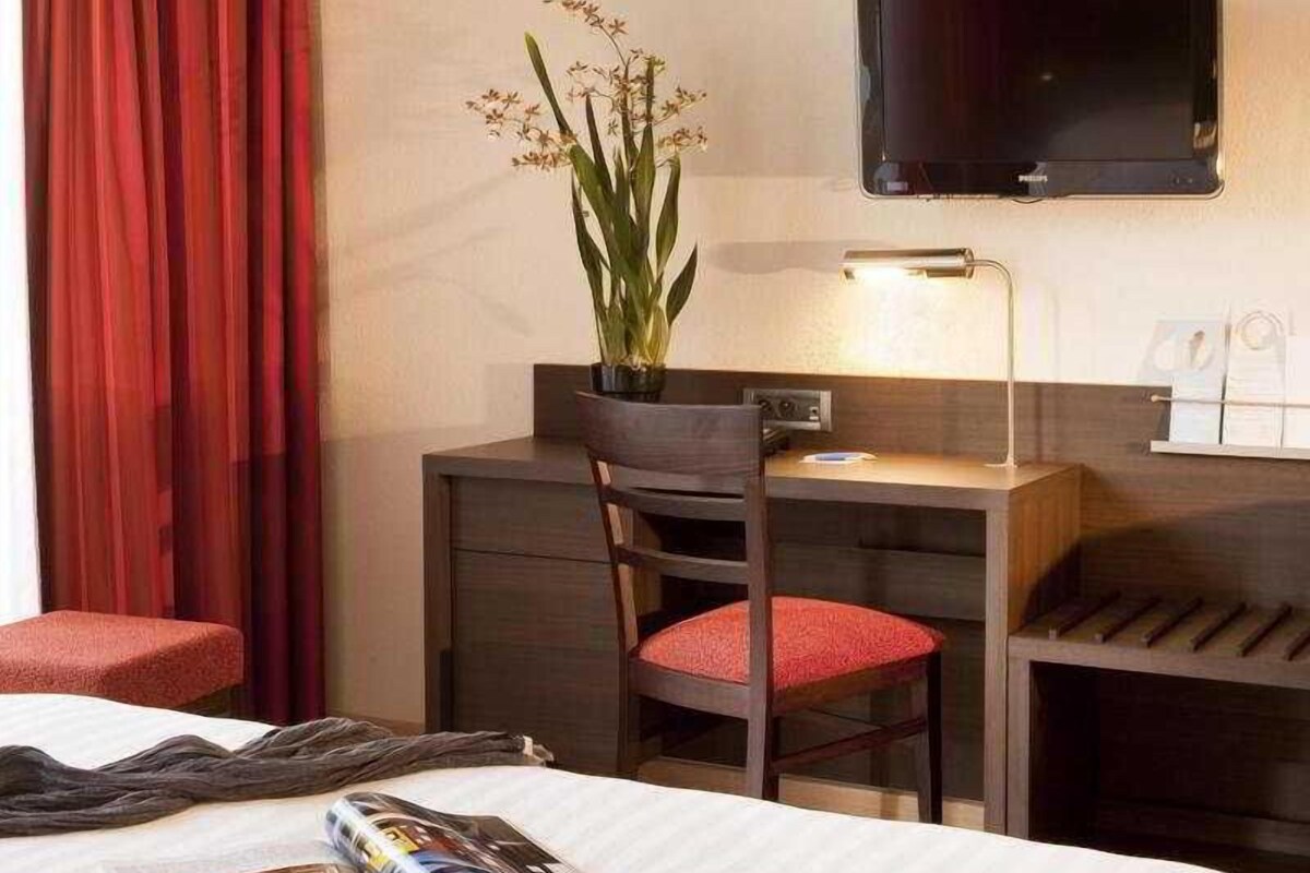 Una habitación de hotel con cama, escritorio, silla, televisión y una planta.