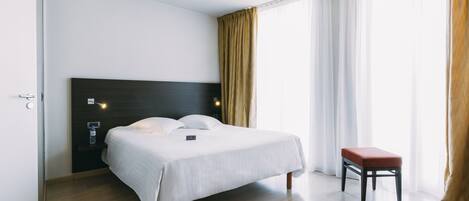 A modern hotel room with a large bed, a bedside table, a small stool, and a large window with curtains.