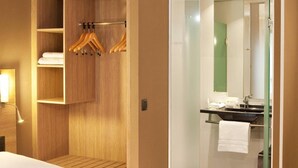Habitación de hotel con cama, armario de madera con ropa colgada, baño con cabina de ducha de cristal y espejo.