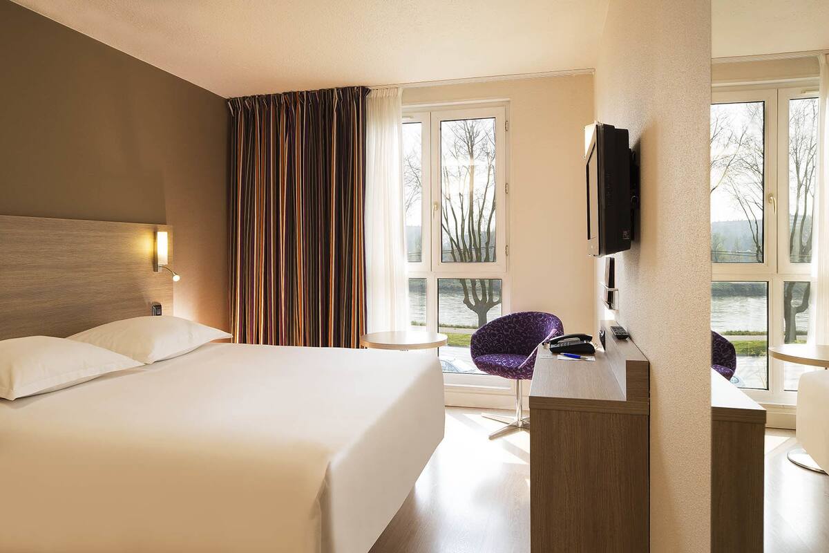 A hotel room with a bed, a desk with a chair, a TV, and large windows with a view of trees and a body of water.