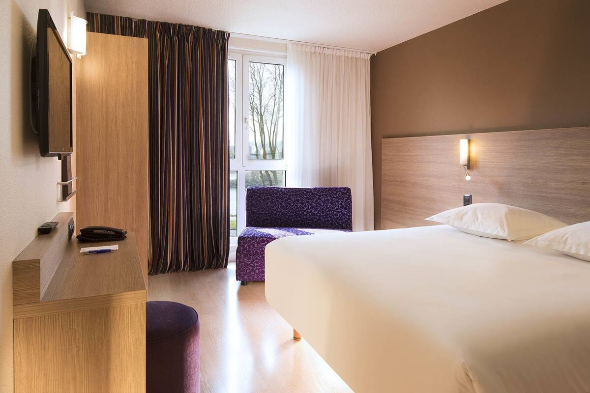 A hotel room with a bed, a desk with a telephone, a chair, and a window with curtains.