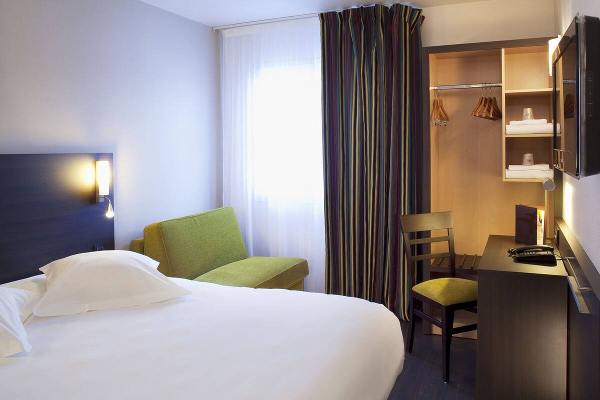 A hotel room with a bed, a desk, a chair, a wardrobe, and a television.