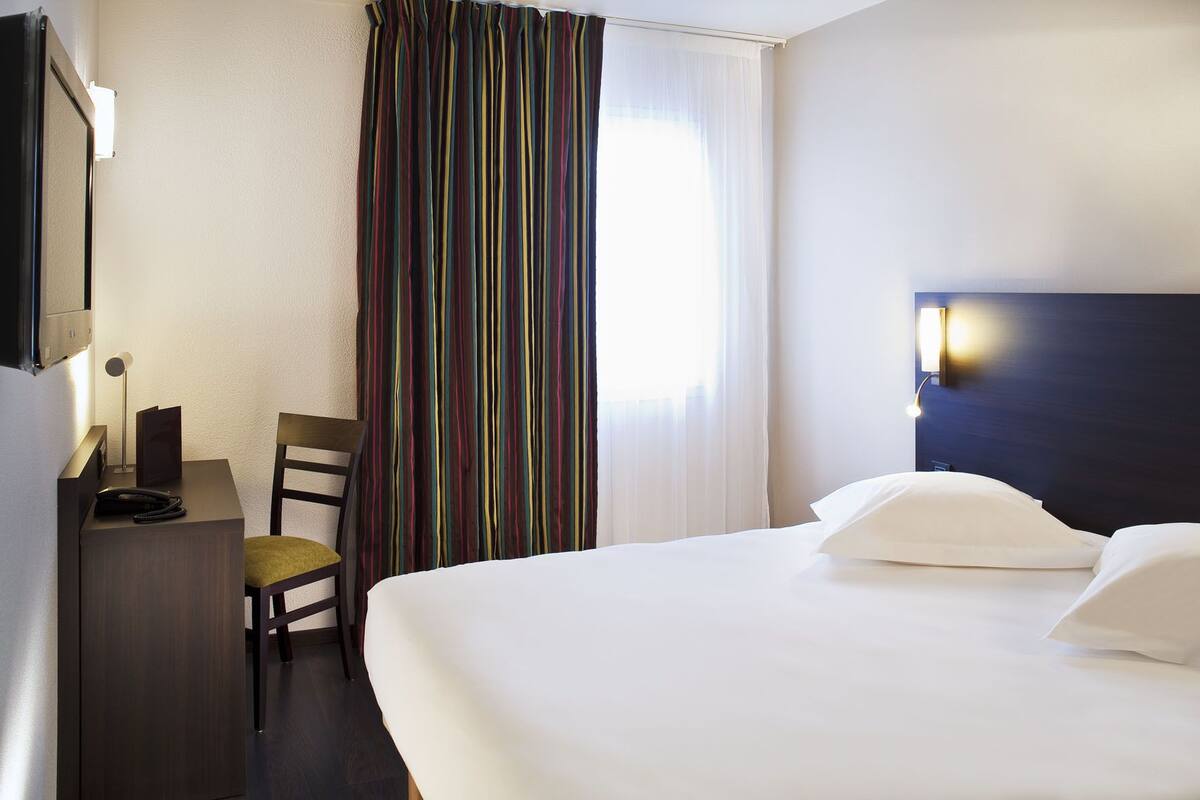 A hotel room with a bed, a desk with a telephone, a chair, a TV, and a window with curtains.