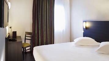 A hotel room with a bed, a desk with a telephone, a chair, a TV, and a window with curtains.