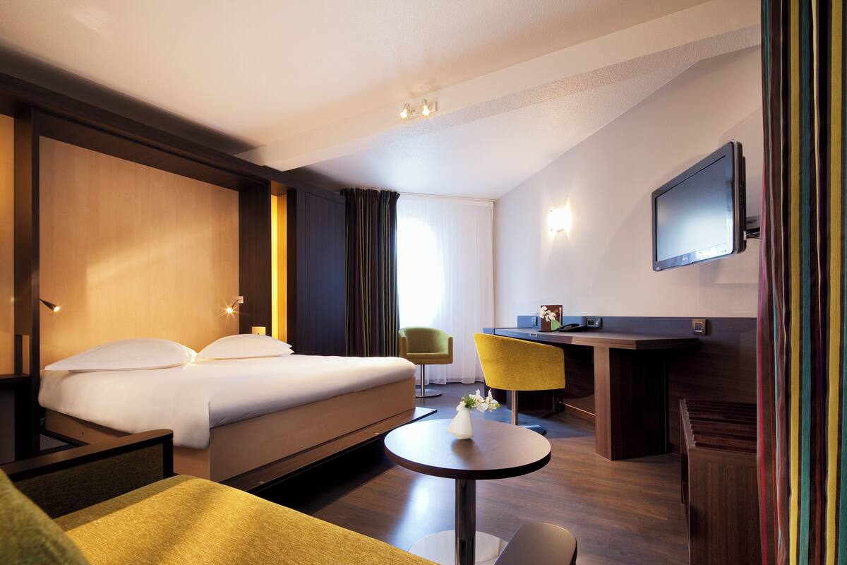 A hotel room with a bed, a desk, a chair, a TV, and a small table with a vase.