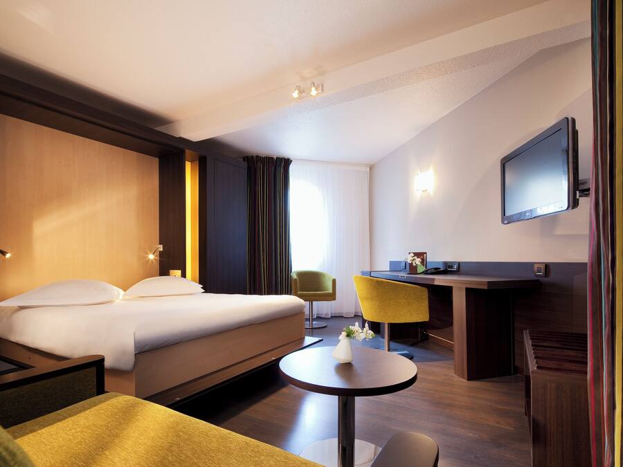 Quarto de hotel com cama, escrivaninha, cadeira, televisão e mesinha de centro com um vaso.