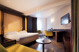 A hotel room with a bed, a desk, a chair, a TV, and a small table with a vase.