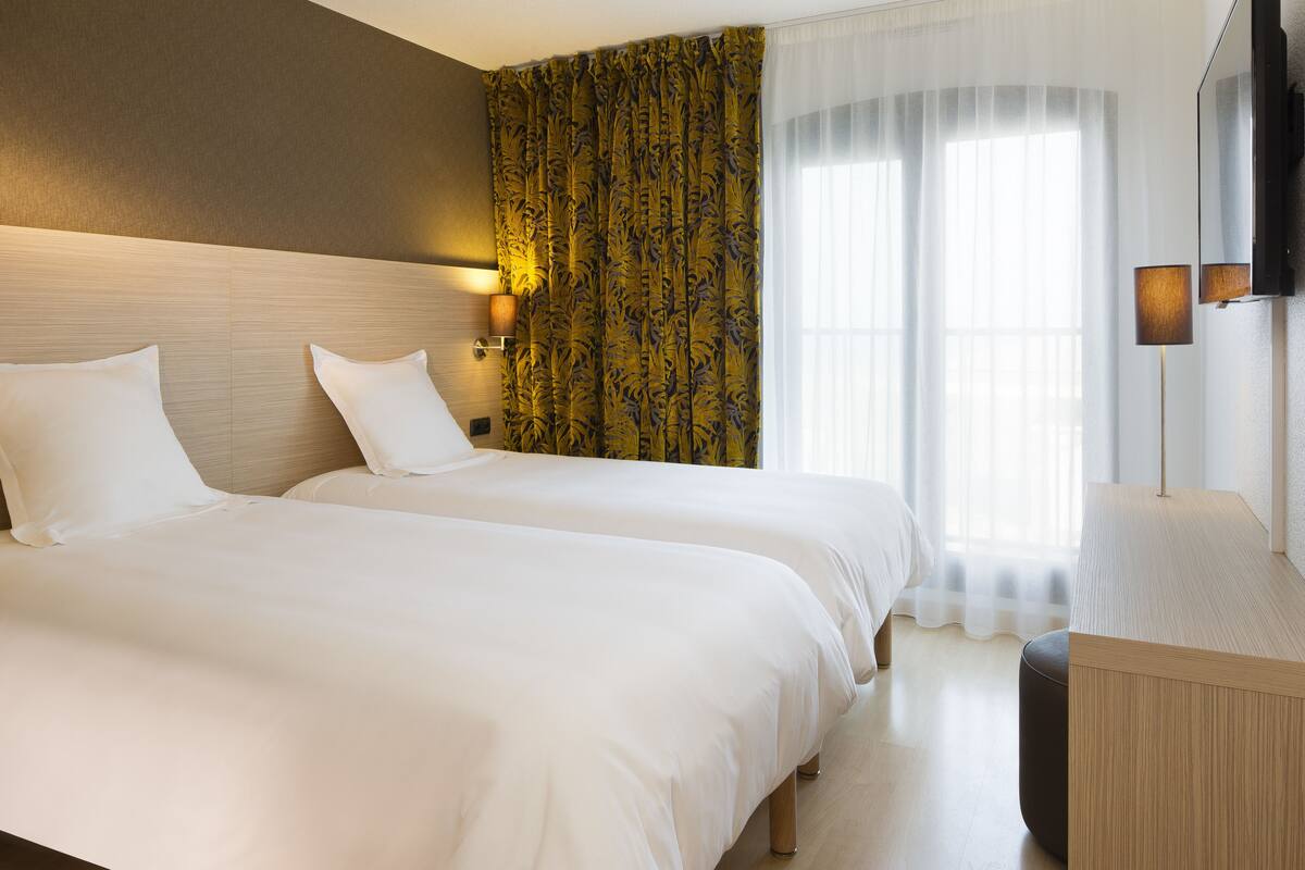 Een hotelkamer met twee bedden, een groot raam met dunne gordijnen, een flatscreen televisie en een bedlamp.