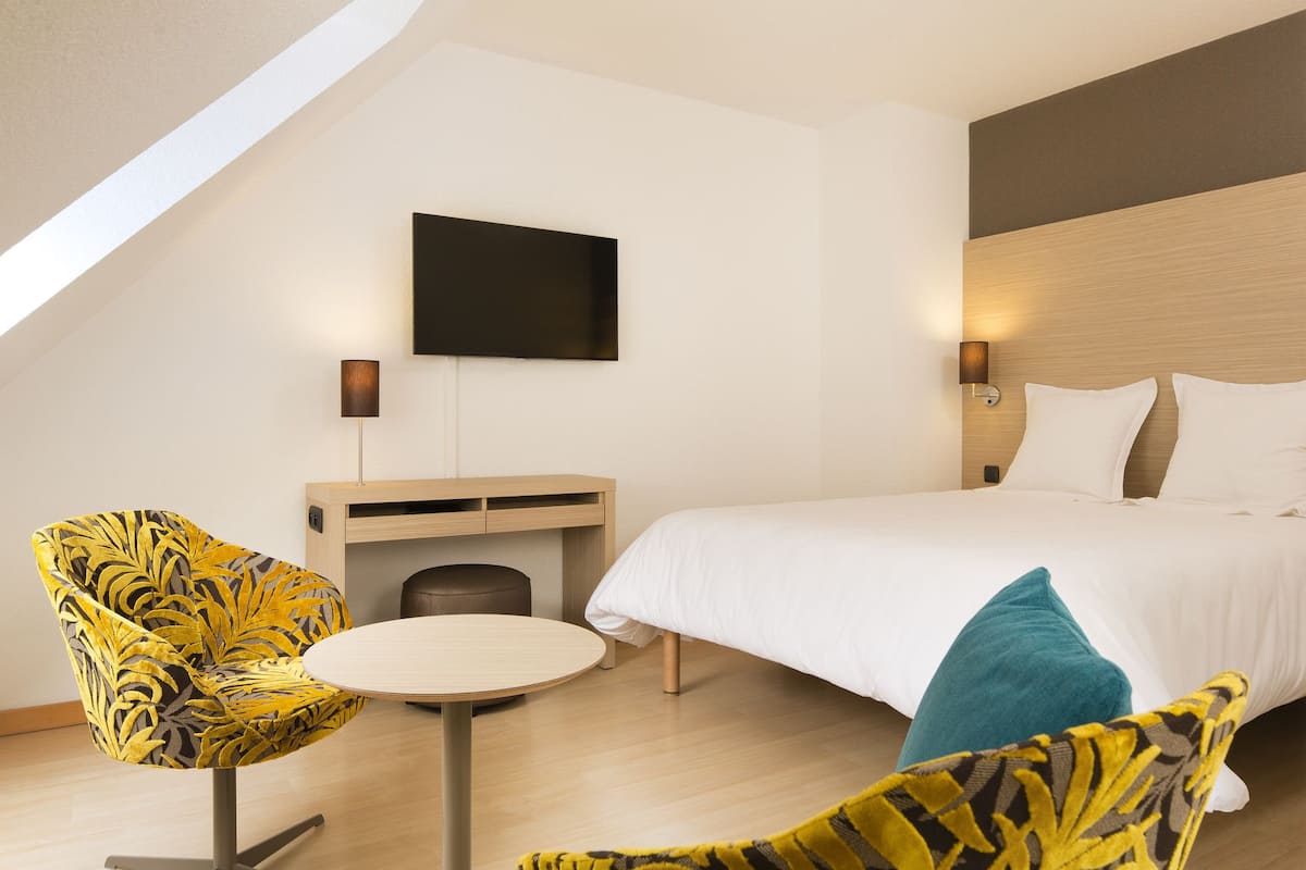Een hotelkamer met een bed, een aan de muur bevestigde televisie, een klein tafeltje en een stoel met bladmotief.