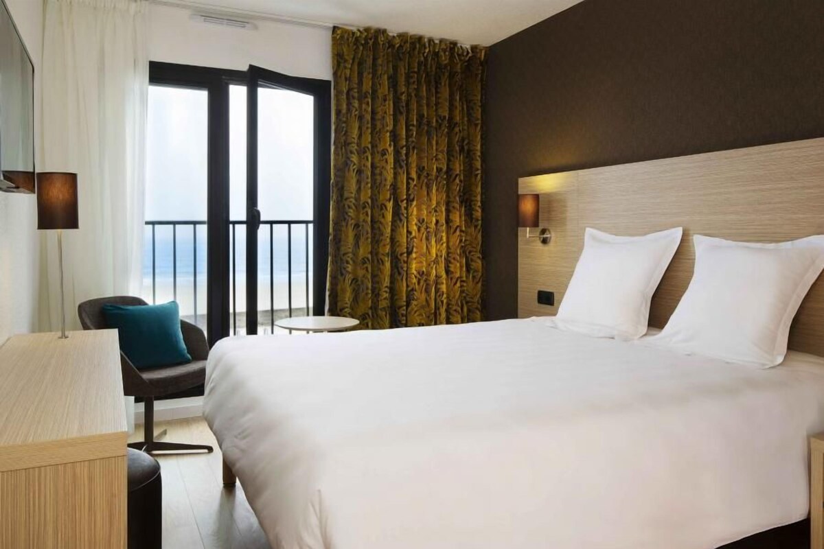 Een hotelkamer met een groot bed, een klein rond tafeltje, een stoel en uitzicht op het strand door een schuifdeur.