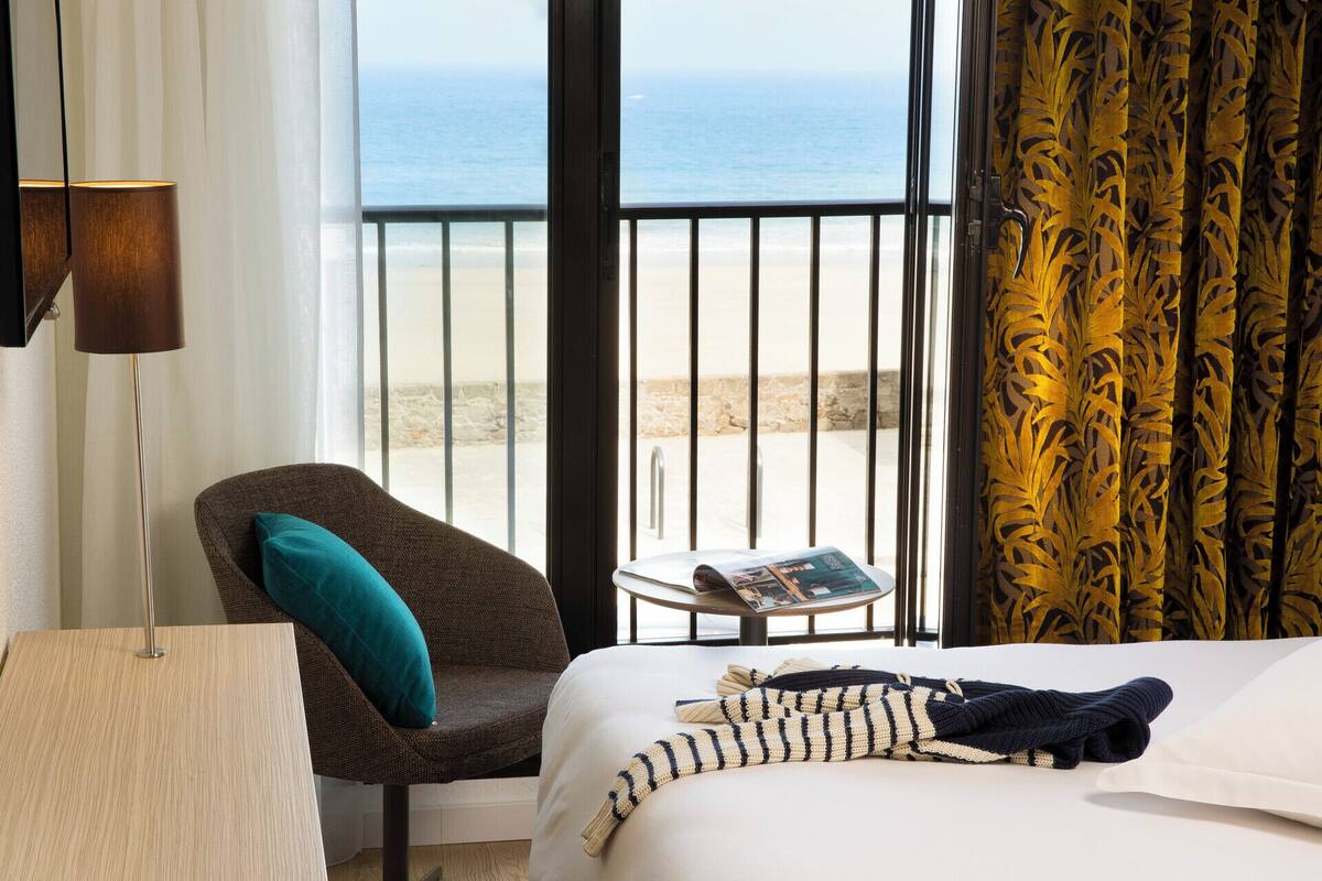 Habitación de hotel con cama, una silla, una mesita con una revista y vistas al océano a través de amplias ventanas.