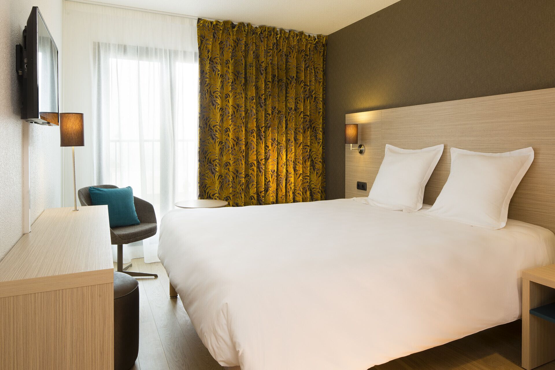 A hotel room with a large bed, a flat-screen TV mounted on the wall, a desk with a lamp, and a chair.