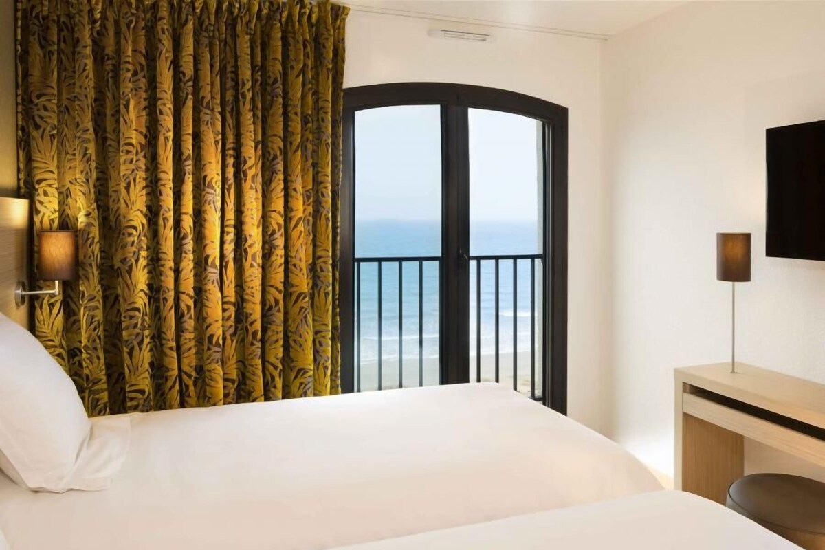 Ein Hotelzimmer mit Bett, Schreibtisch, Lampe, Fernseher und einem Balkon mit Meerblick.