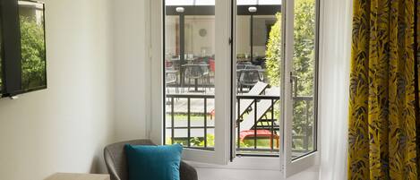 A hotel room with a bed, a chair with a pillow, a TV on the wall, and a view of the outdoor seating area through the glass doors.