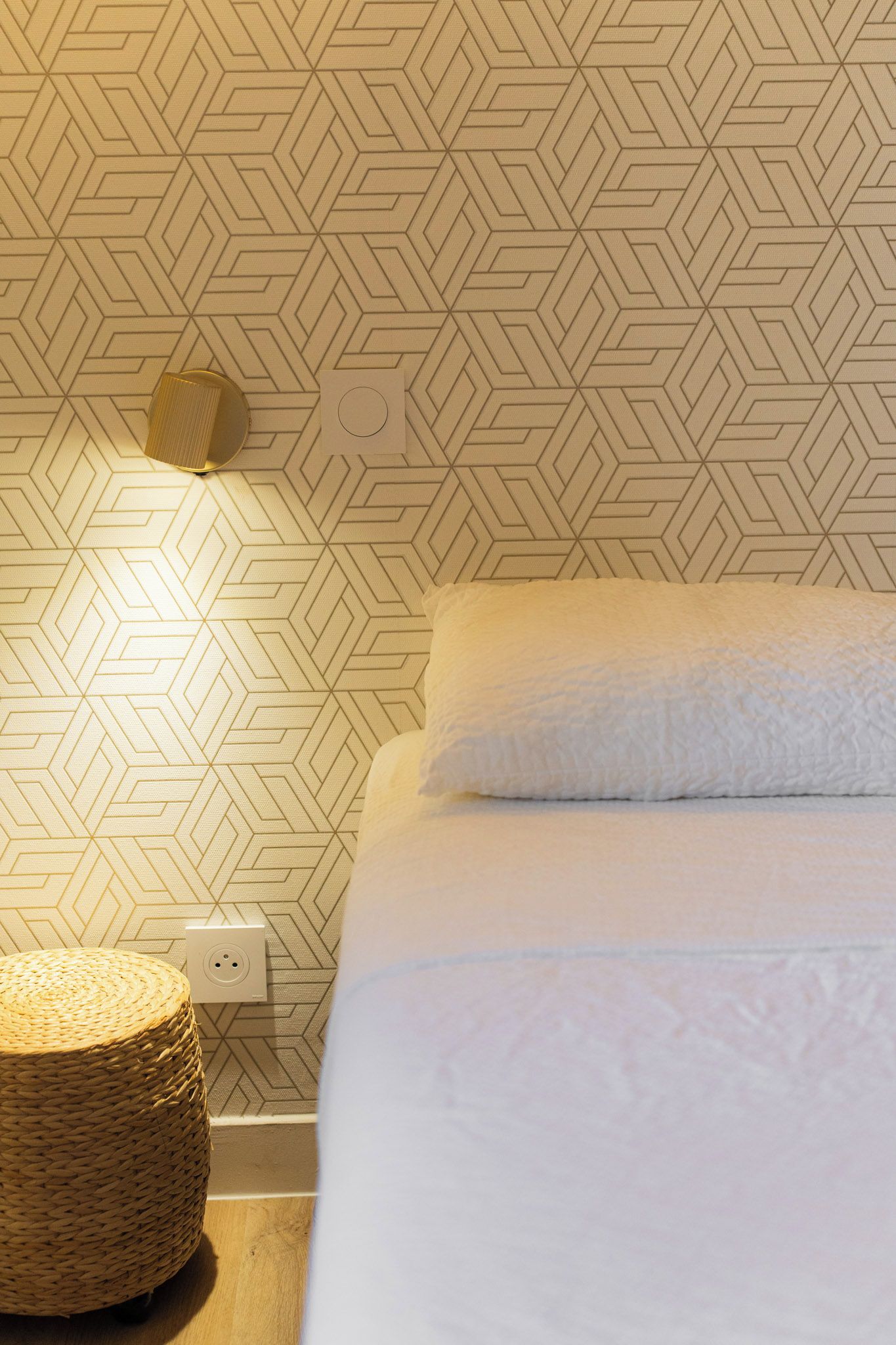 Ein Zimmer mit einer Wand mit geometrischem Muster, einem Bett mit weißer Bettwäsche, einer wandmontierten Lampe und einem gewebten Korb.