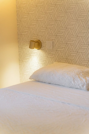 A bed with a textured white cover, a wall with a geometric pattern, and a wall-mounted light fixture.