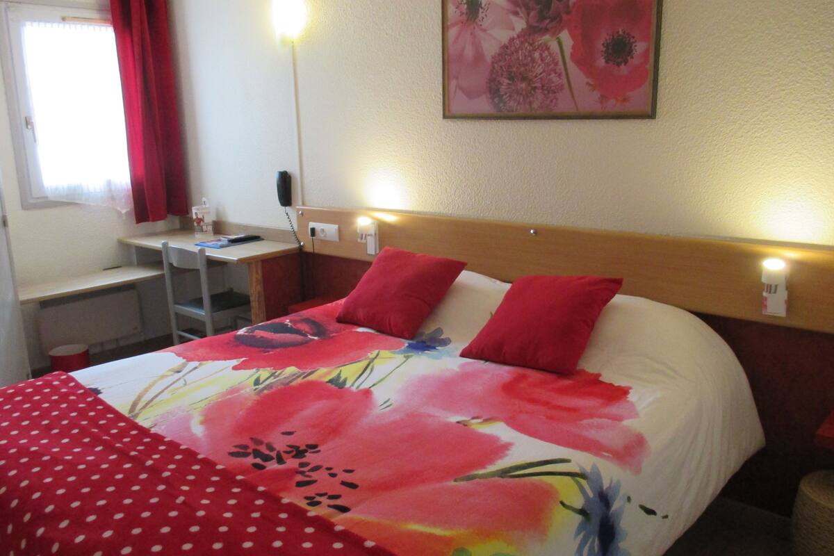 A hotel room with a bed, a desk, and a chair. There is a painting on the wall and a window with curtains.