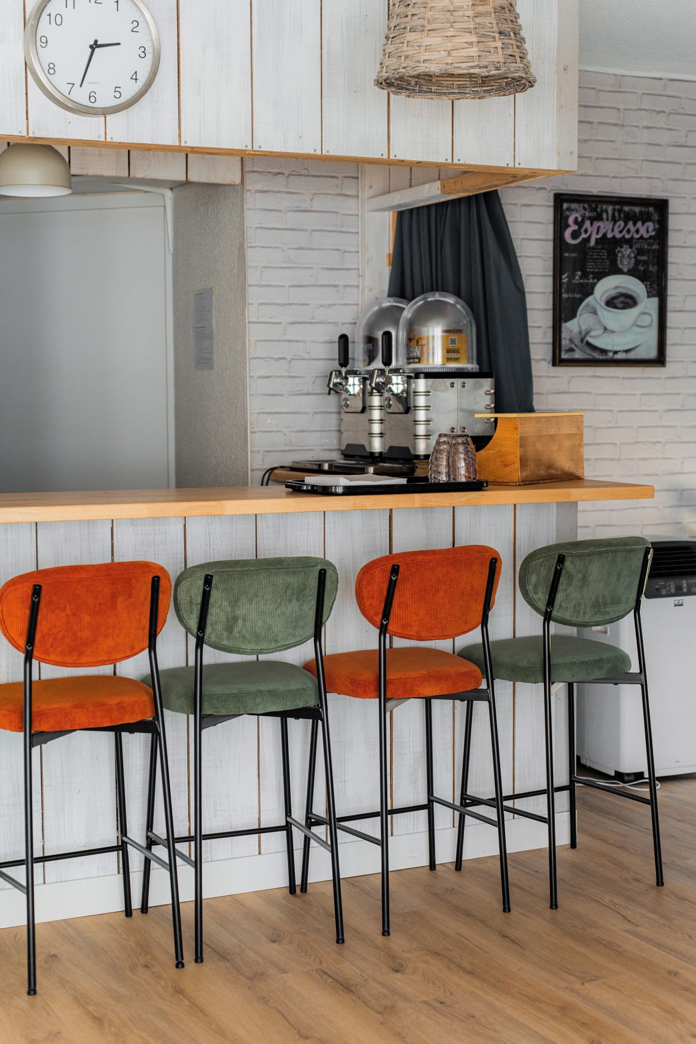 A kitchen with a wooden bar, four bar stools, a clock, a coffee machine, and a poster on the wall.