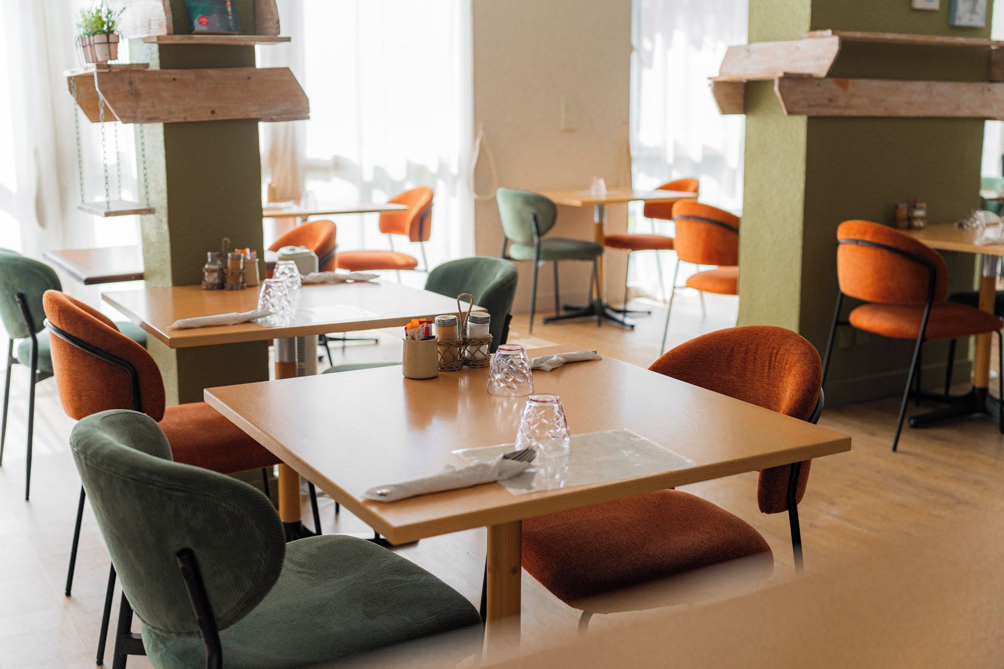 Un espace repas moderne avec des tables et des chaises en bois, dont certaines sont dotées de coussins. Des plantes en pot sont disposées sur des étagères, et l’espace est bien éclairé.