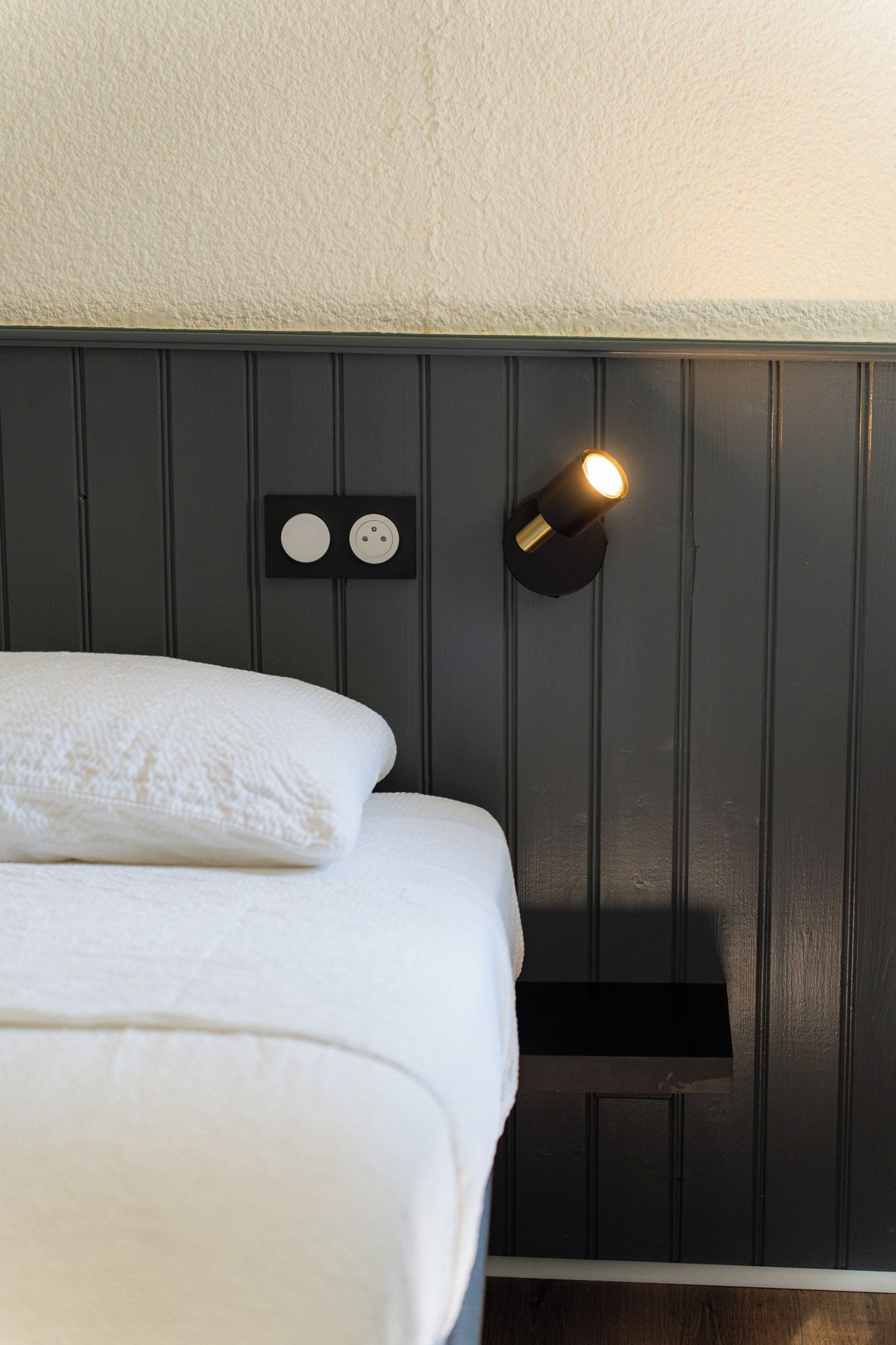 En seng med hvide sengetøj, en vægmonteret lampe og en lille hylde.