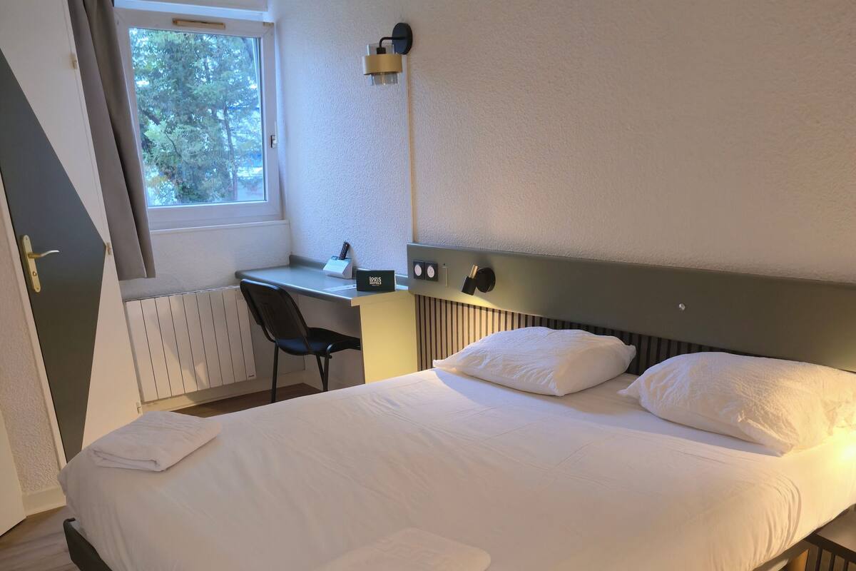 A neatly made bed with white linens and two pillows, a desk with a chair, a lamp, and a window with curtains.