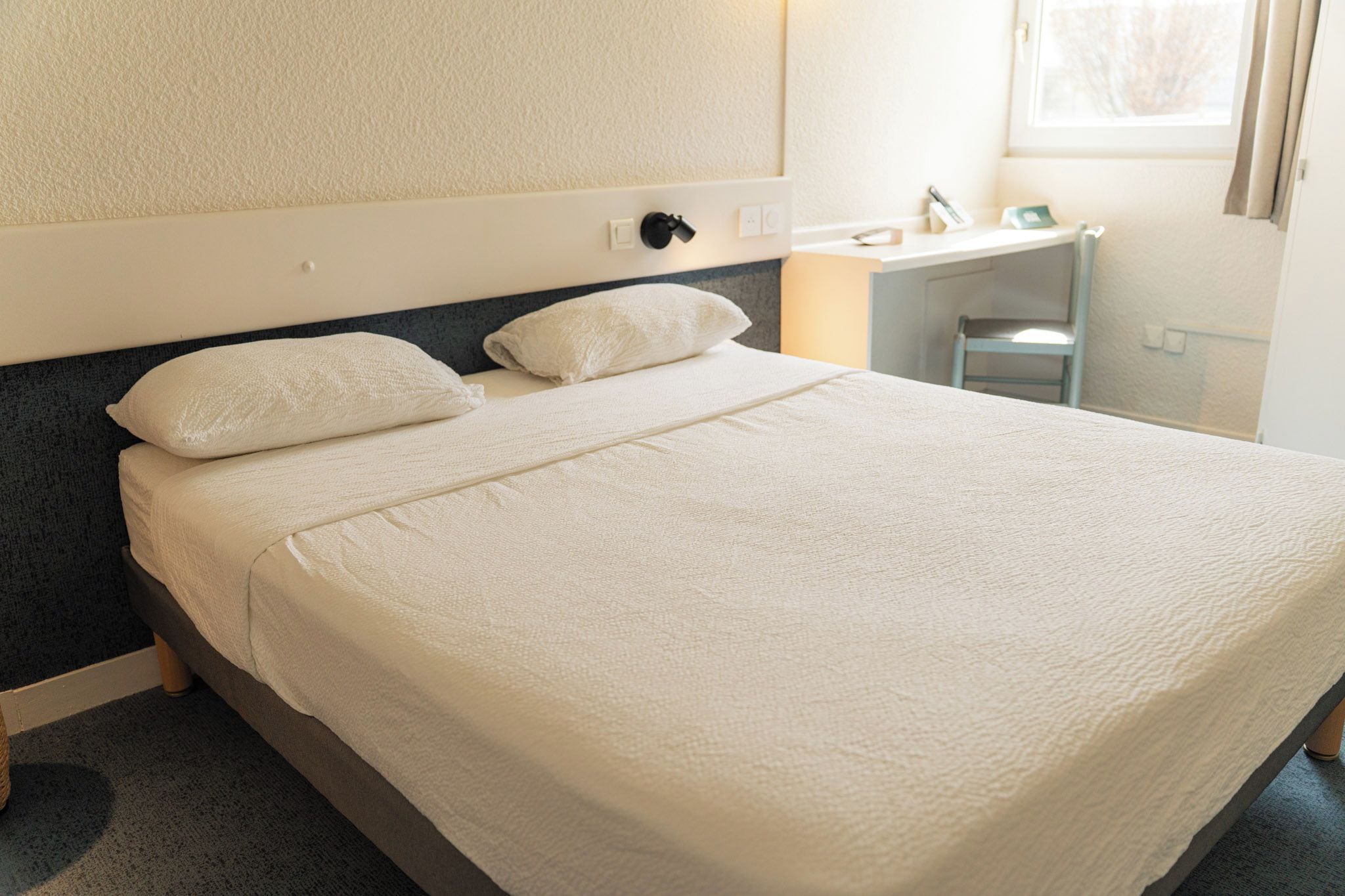 Un lit bien fait, avec une couette et des oreillers blancs, une tête de lit sombre et une table de chevet sur laquelle se trouve une lampe.