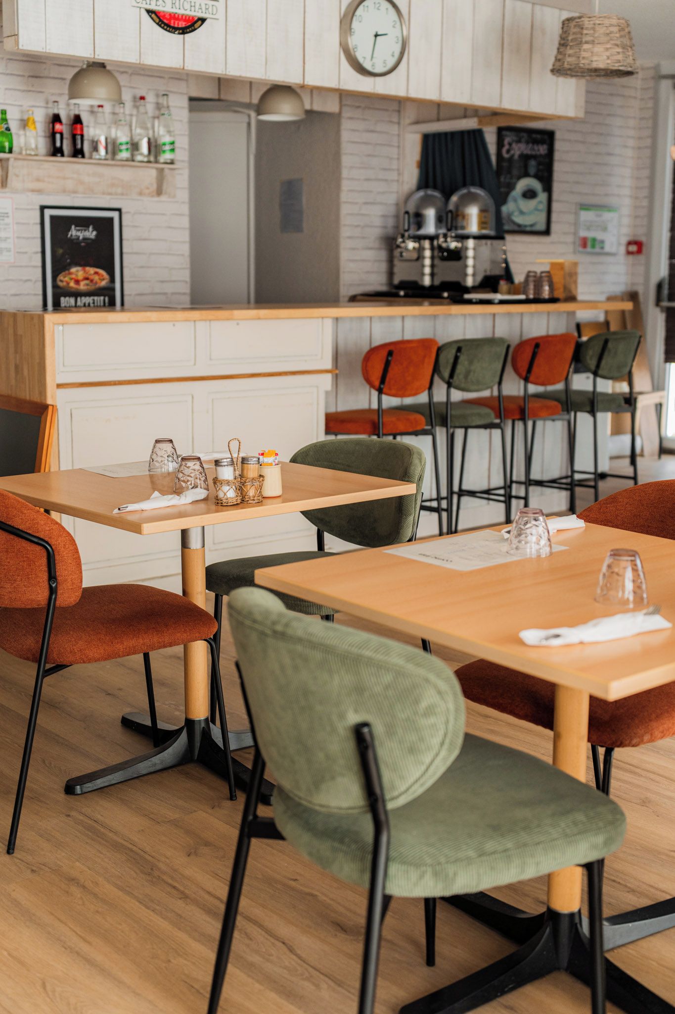 L’intérieur d’un restaurant, avec des tables et des chaises en bois, un espace cuisine et une horloge au mur.