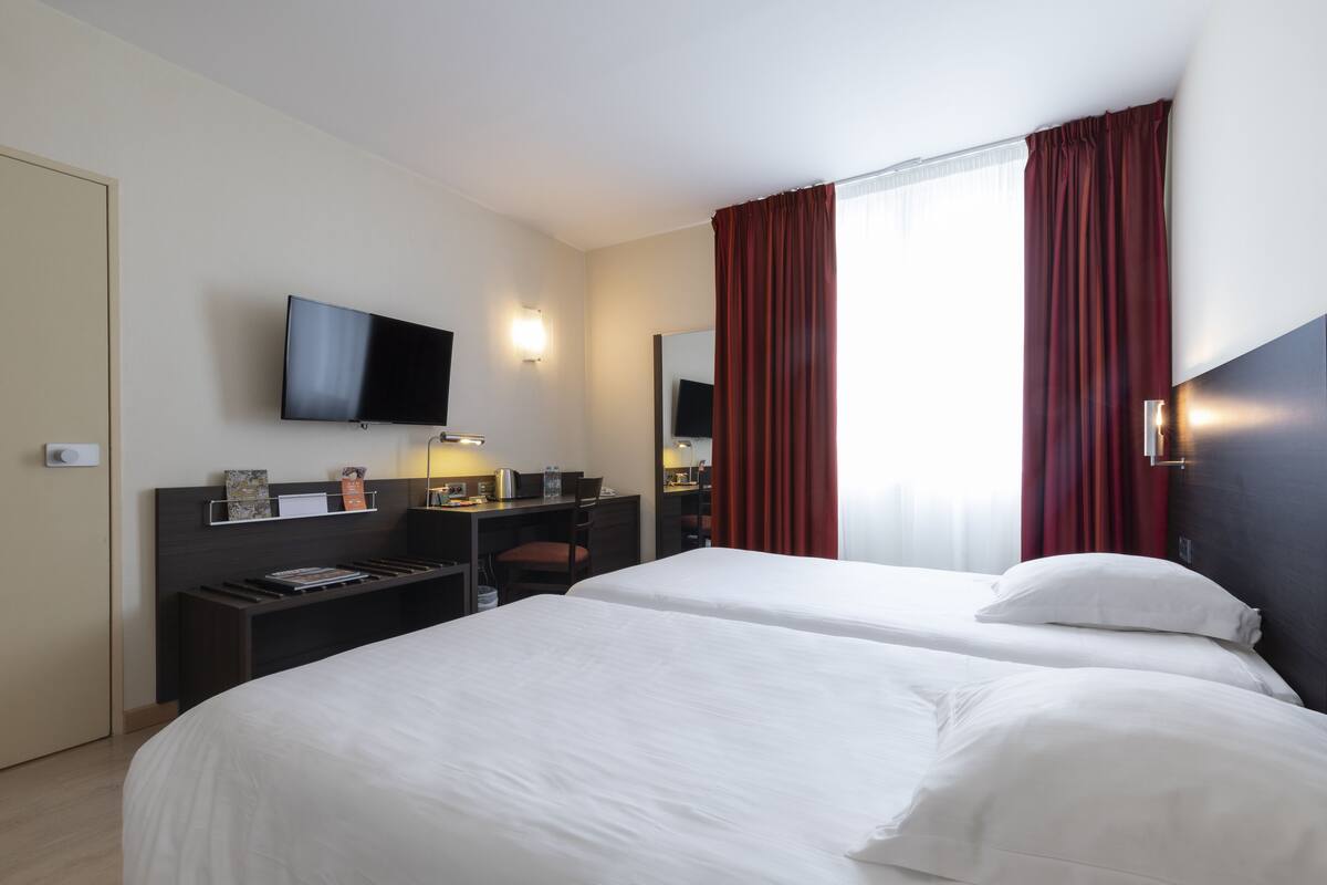 Habitación de hotel con dos camas, un escritorio, un televisor de pantalla plana y un ventanal con cortinas rojas.