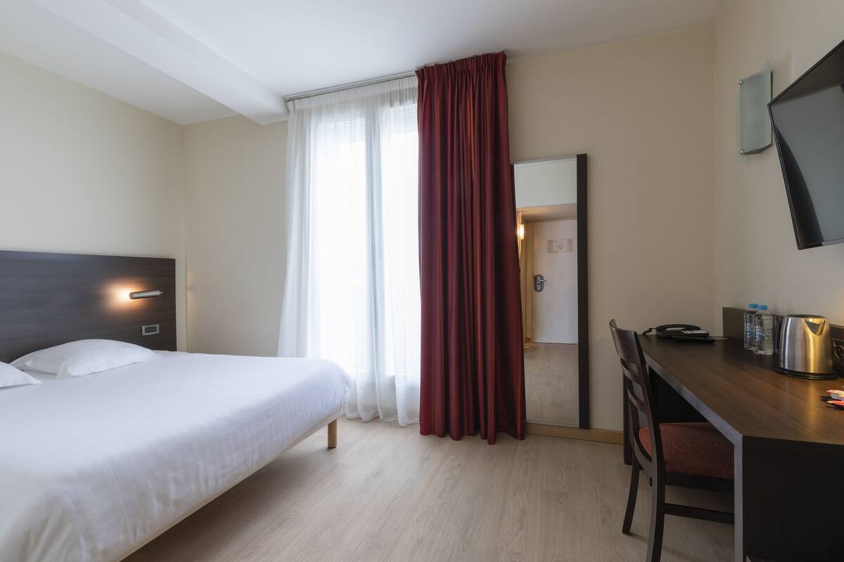 Habitación de hotel con cama, escritorio, silla, espejo y ventana con cortinas.