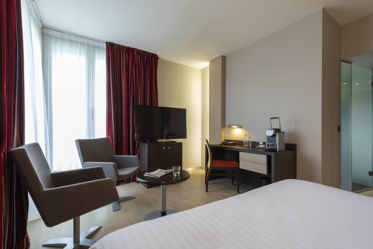 Habitación de hotel con cama, dos sillones, escritorio con computadora, televisor y una mesita con revista y vaso.