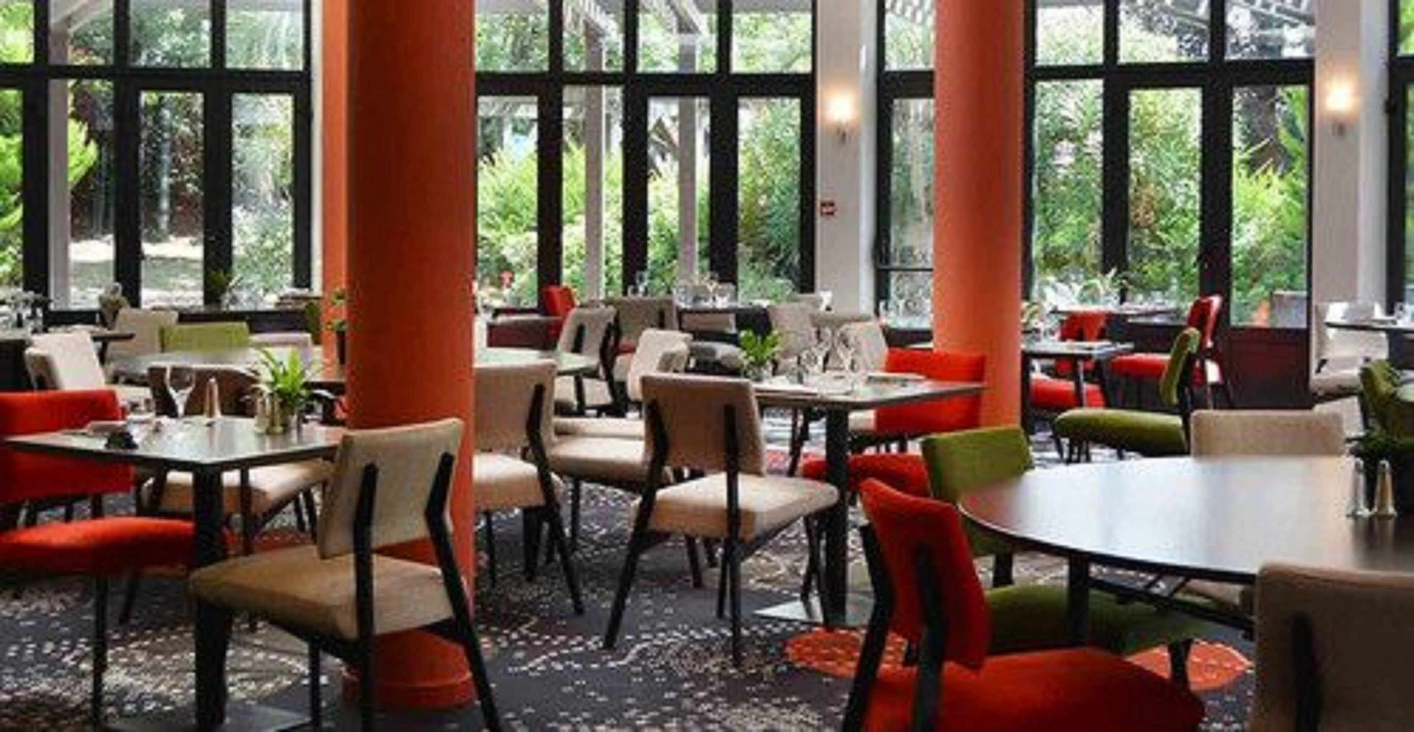Un restaurant doté de nombreuses tables et chaises, de grandes fenêtres et de plantes d’intérieur.