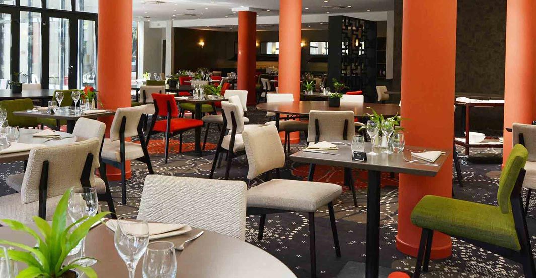 Un comedor de hotel con varias mesas y sillas, ventanas amplias y suelo alfombrado.