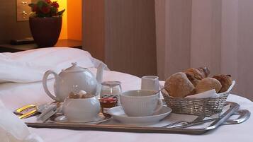A neatly arranged breakfast tray with a teapot, cups, a small bowl of cereal, a basket of bread, and a teapot lid on a bed.