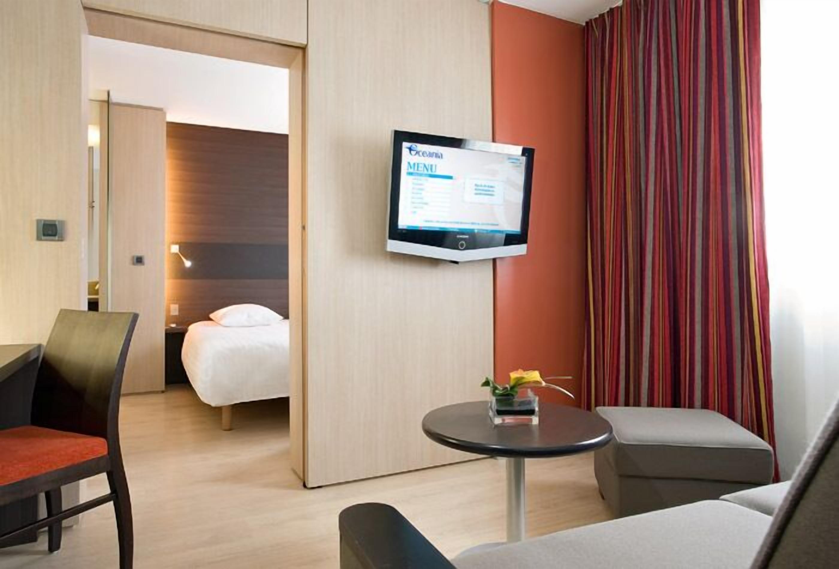 A hotel room with a bed, a desk, a chair, a small table with a flower vase, and a TV displaying a menu.