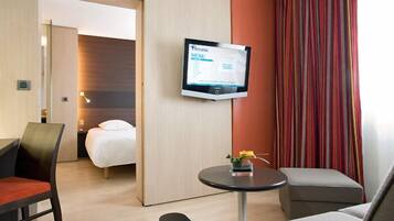 A hotel room with a bed, a desk, a chair, a small table with a flower vase, and a TV displaying a menu.