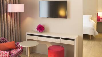 Een moderne hotelkamer met een wandtelevisie, een klein bureau met lades, een ronde tafel, een rode, cilindervormige poef, een gestreept stoel met oranje kussen, en een bed met wit beddengoed.
