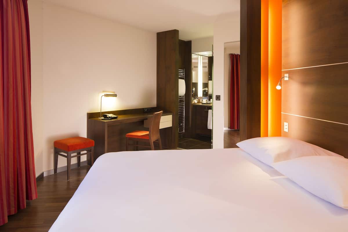 A hotel room with a large bed, a desk with a chair, and a mirror.