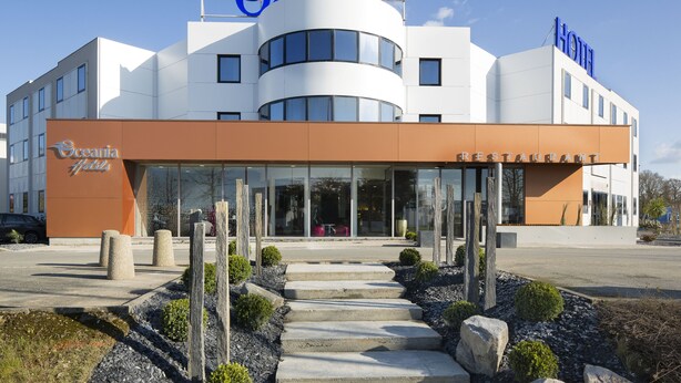 A modern hotel with a restaurant on the ground floor, featuring a curved facade, large windows, and a prominent logo.