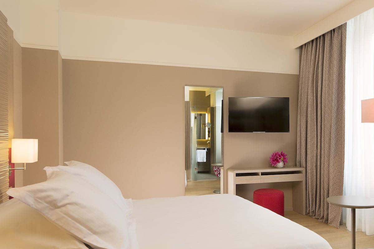 A hotel room with a large bed, a desk with a flat-screen TV, a mirror, and a small table.
