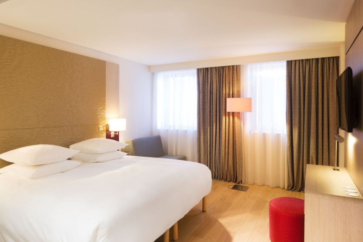 A hotel room with a bed, a desk, a chair, a TV, and a window with curtains.